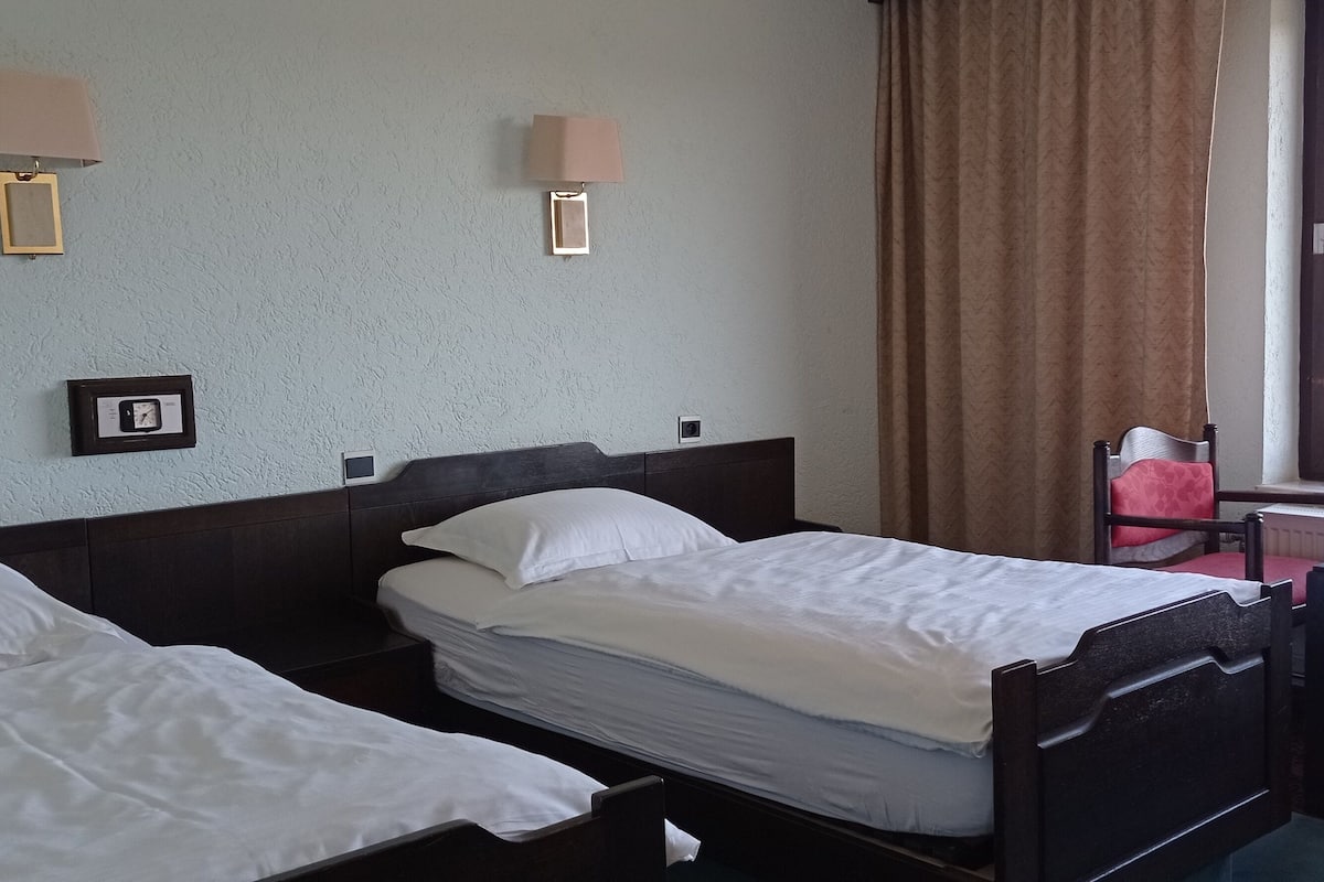 A hotel room with two single beds, a small table, a chair, a window with curtains, and two wall-mounted lamps.