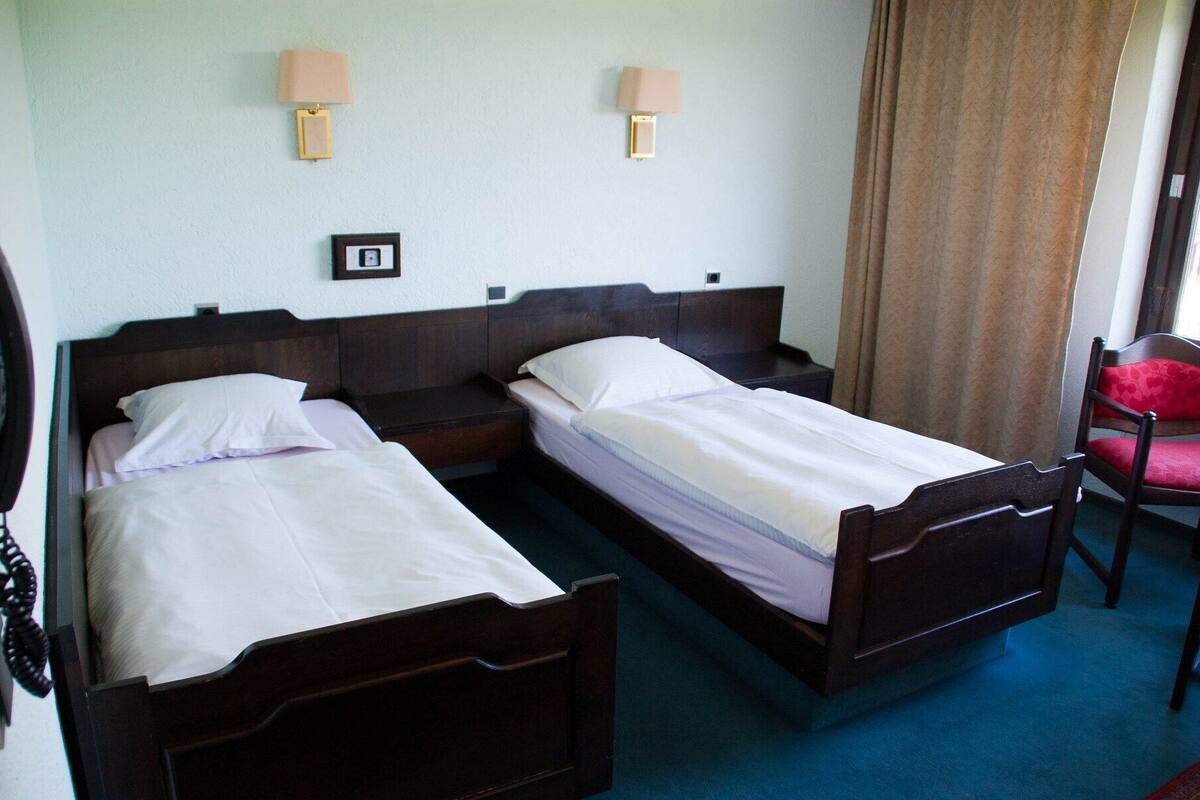 A hotel room with two single beds, a wooden headboard, a bedside table, a lamp, and a chair.