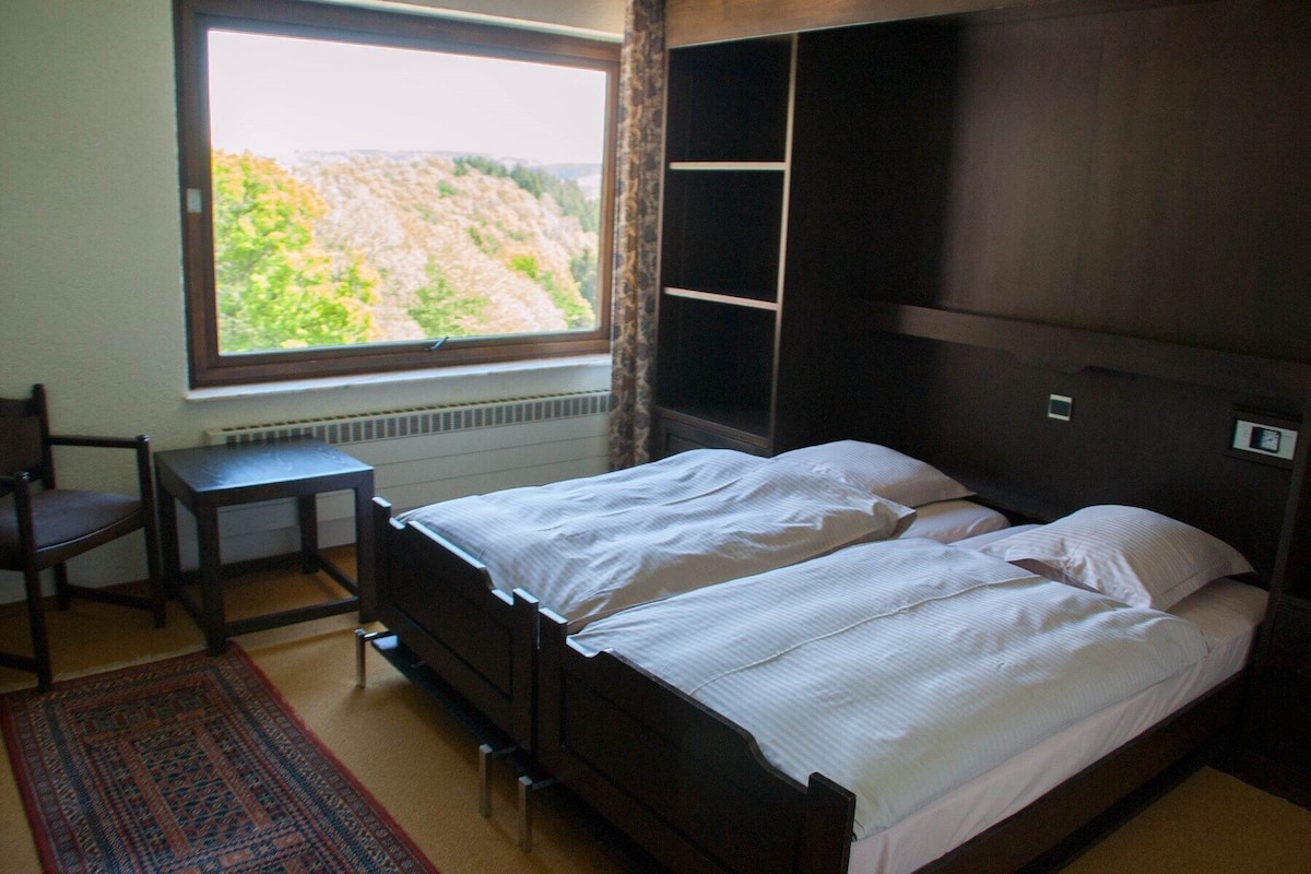 A bedroom with a bed, a chair, a desk, and a window with a view of greenery.