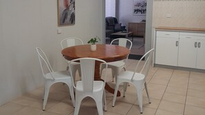 Un área de comedor con una mesa redonda de madera rodeada de sillas blancas, una pequeña planta en maceta y dos cuadros en las paredes.