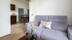 A living room with a grey sofa, a wooden coffee table, and a vase with flowers. There is a dining area in the background with a table and chairs.