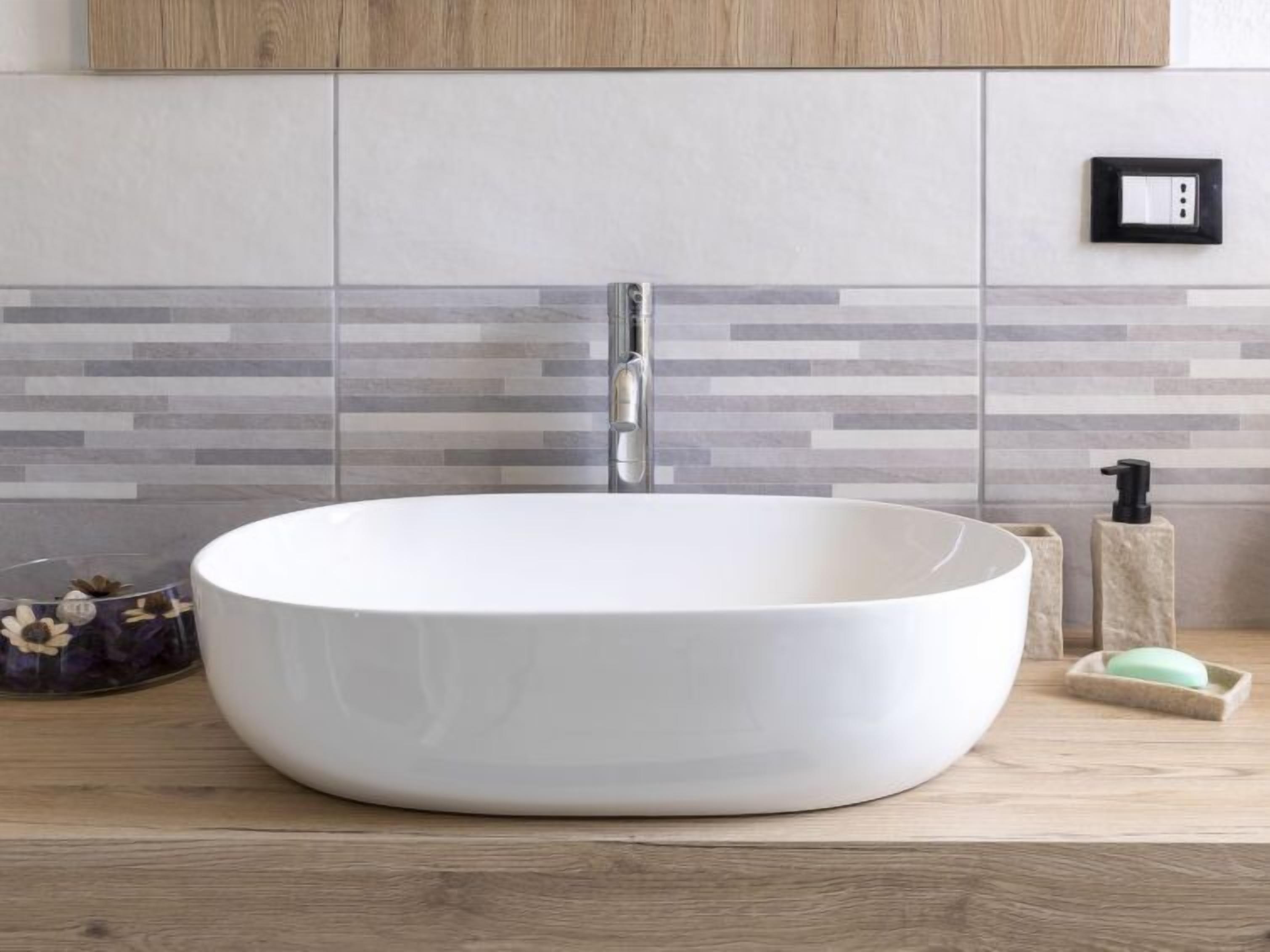 A modern bathroom with a large white vessel sink, a wooden countertop, a soap dispenser, and a wall-mounted light switch.