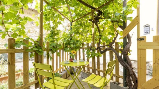 A wooden pergola with green chairs and a table, surrounded by lush vines.