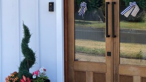 Une porte à double battant en bois avec des panneaux de verre, deux couronnes décoratives accrochées à la porte, une plante en pot placée à côté de la porte et un luminaire fixé au mur.