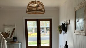 Une cuisine avec une table en bois, deux tabourets, un luminaire suspendu et une vue sur l’extérieur par une porte.