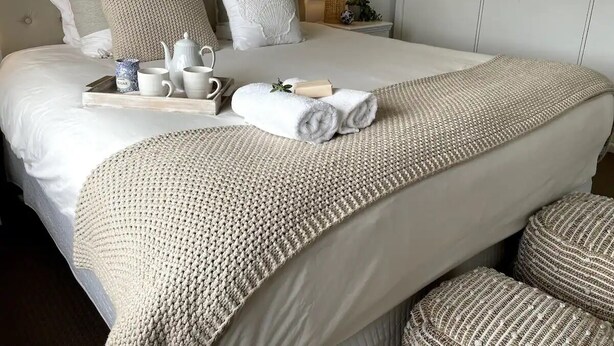 Un lit bien fait, avec des draps blancs, un plaid beige en tricot et des serviettes enroulées.