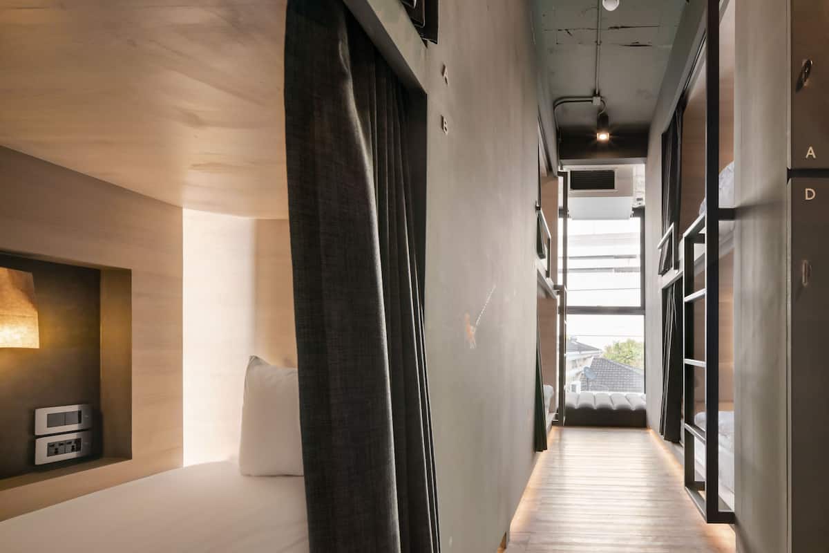 Un estrecho pasillo con suelo de madera, una cama a la izquierda, una repisa con lámpara y una puerta a la derecha.
