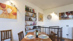 A dining area with a wooden table set for four, surrounded by chairs. A bookshelf filled with books and a painting of flowers are on the wall.