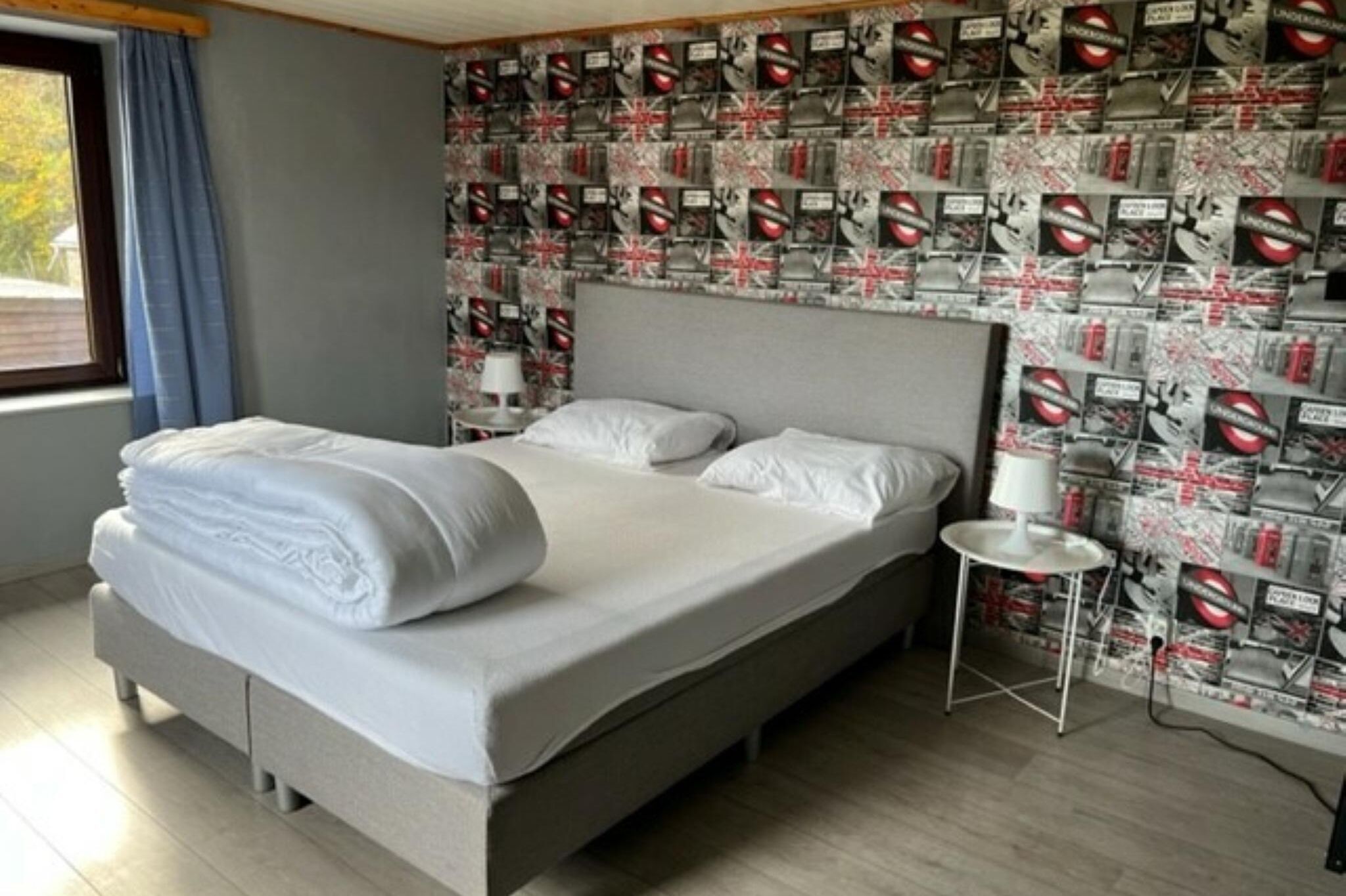 A neatly made bed with white linens, a grey headboard, and a small bedside table with a white lamp. The room features a patterned wallpaper with circular designs and a window with blue curtains.