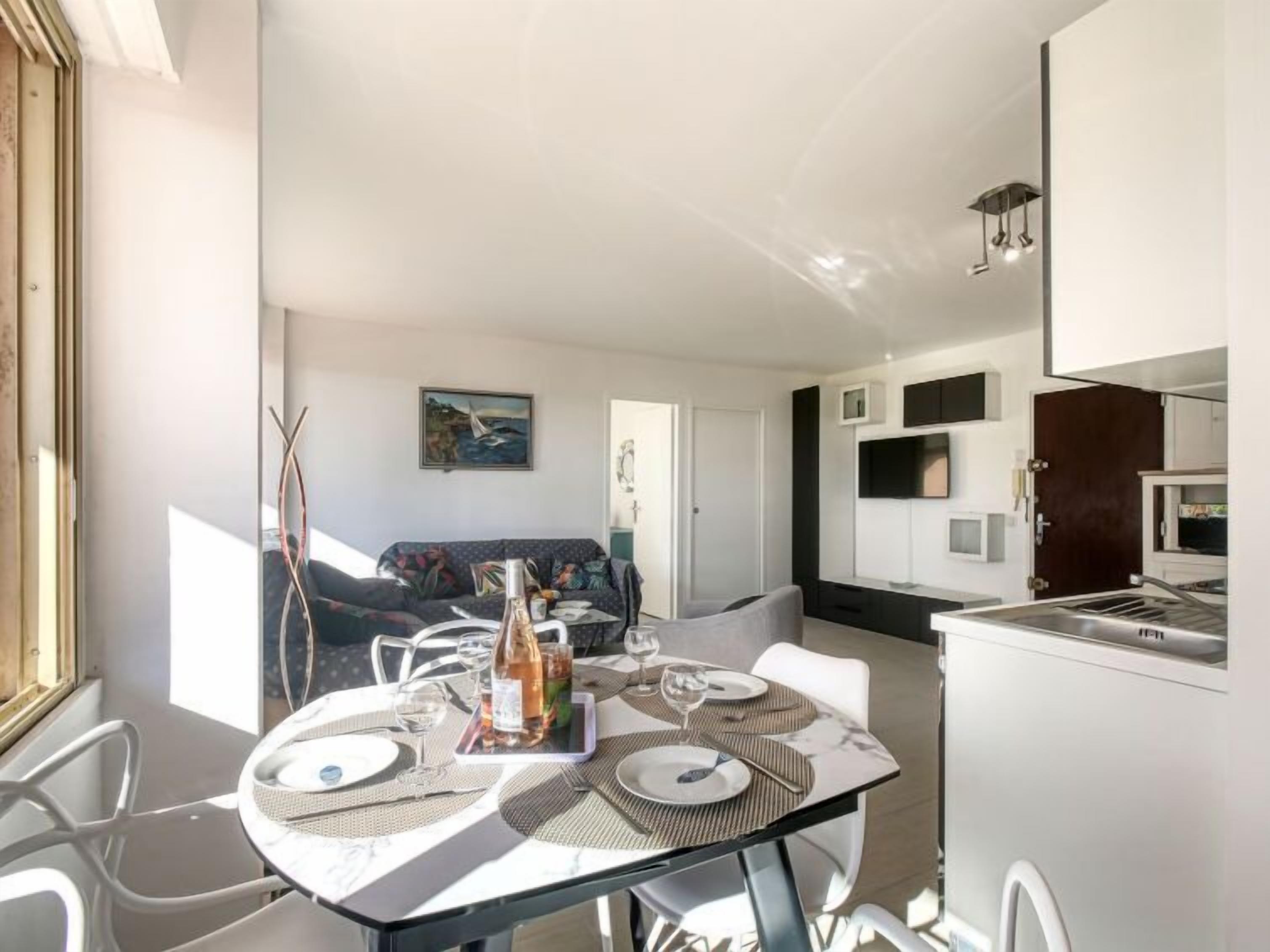 A modern, well-lit dining area with a table set for two, a sofa in the background, and a kitchen area with a TV and cabinets.