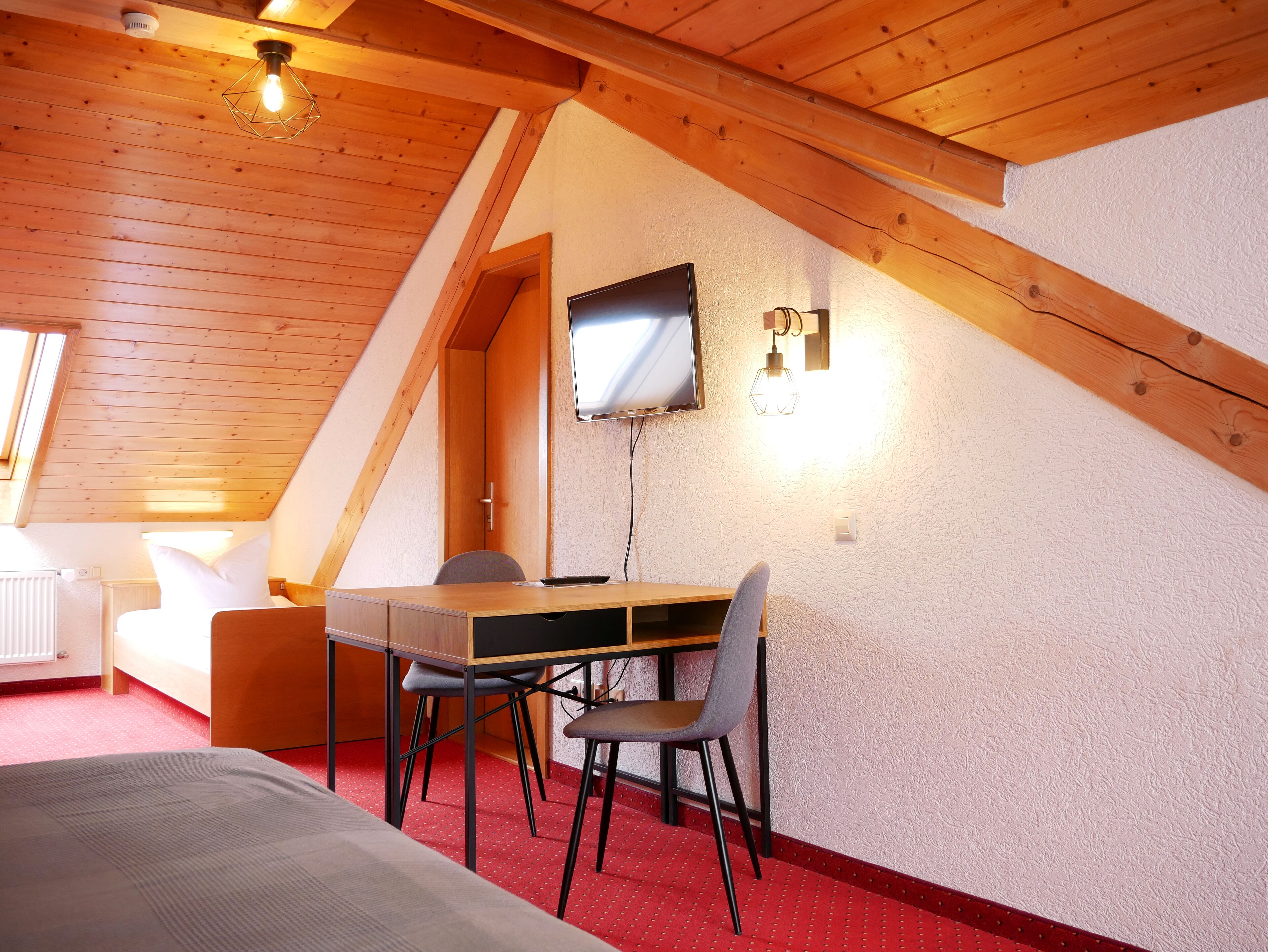 Una habitación acogedora con techo inclinado, un escritorio de madera, una silla, una cama y un televisor en la pared.