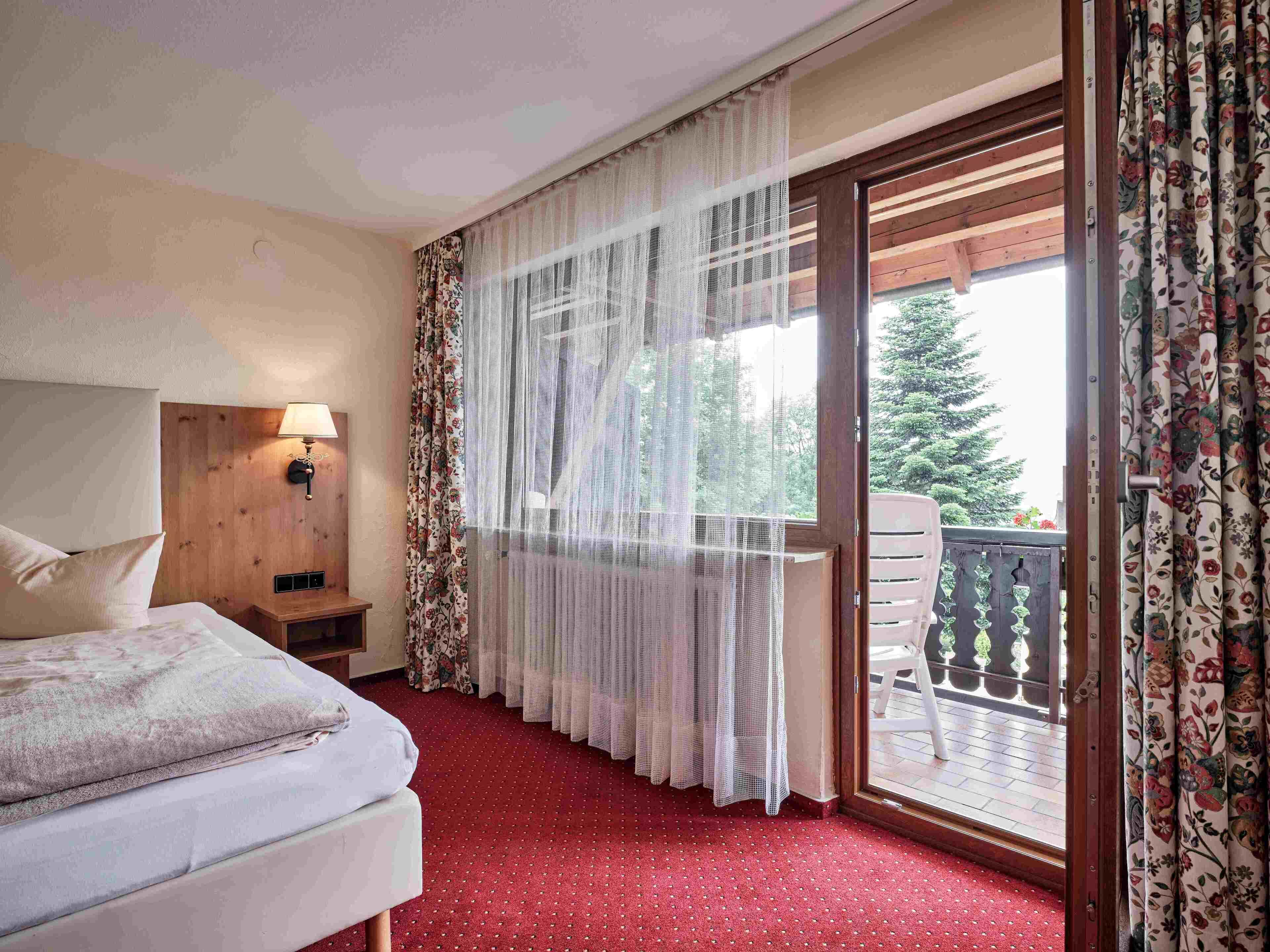 Habitación de hotel con cama, mesita de noche, lámpara, balcón con vista a árboles y alfombra roja.