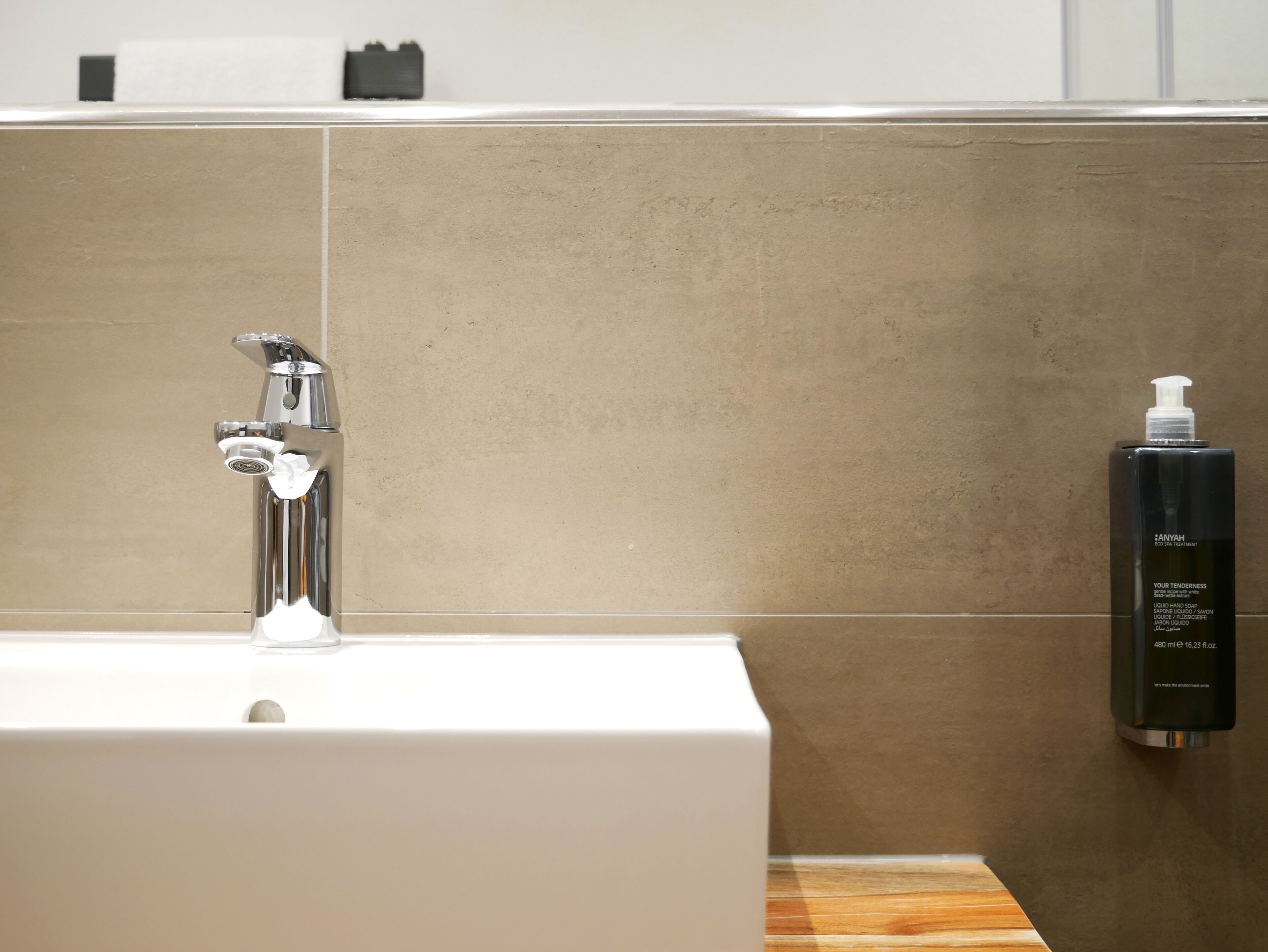 A modern bathroom with a chrome faucet, a white sink, and a black bottle of AnyaH hand soap on a wooden countertop.
