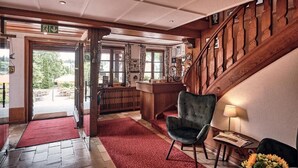 A lobby with a wooden staircase, a reception desk, and a red carpet leading to an outdoor area. - Hotel Hochfirst (Lenzkirch)
