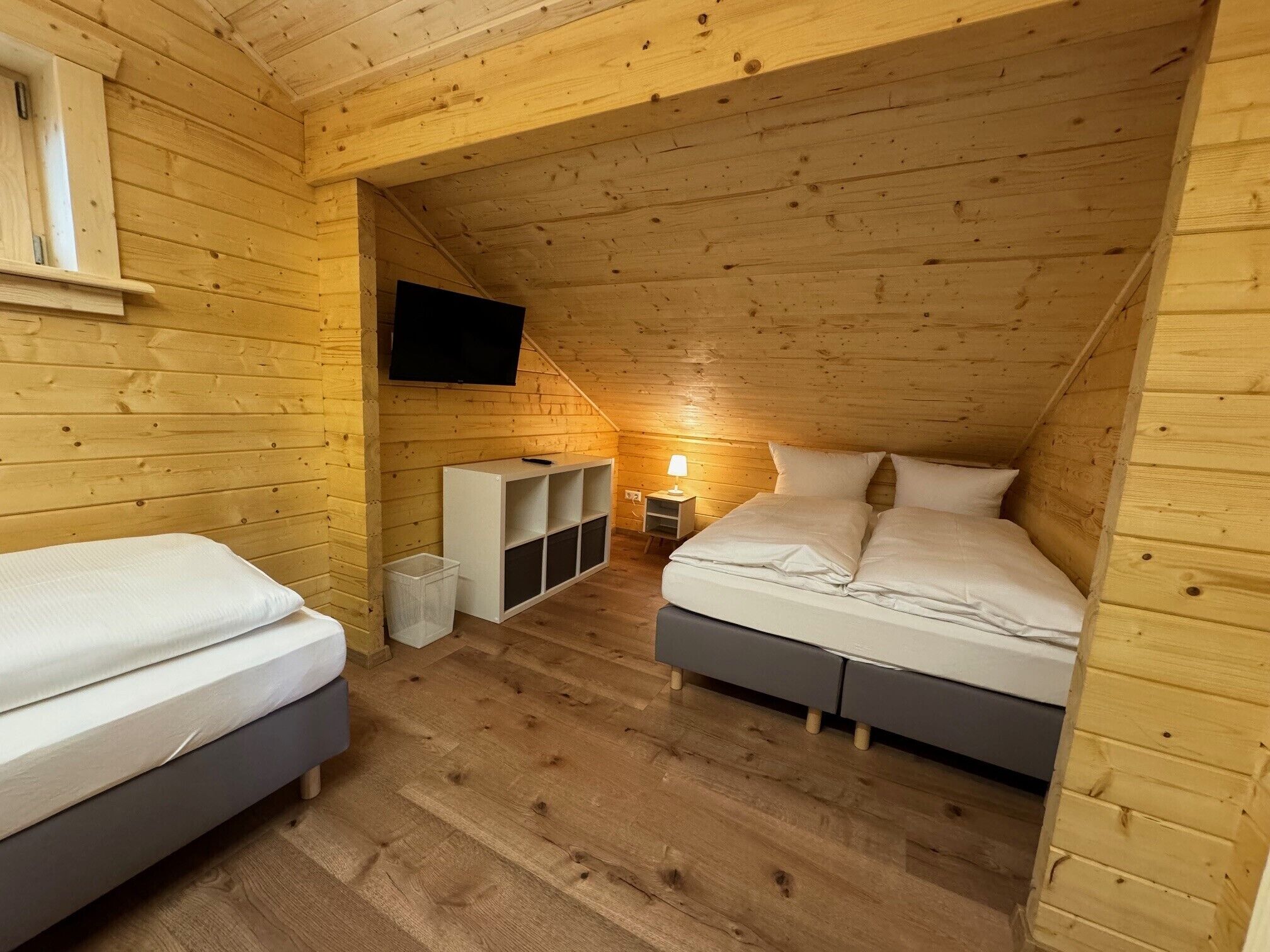 A cozy attic bedroom with wooden walls, a TV mounted on the wall, a bed, a bedside table with a lamp, and built-in shelves.