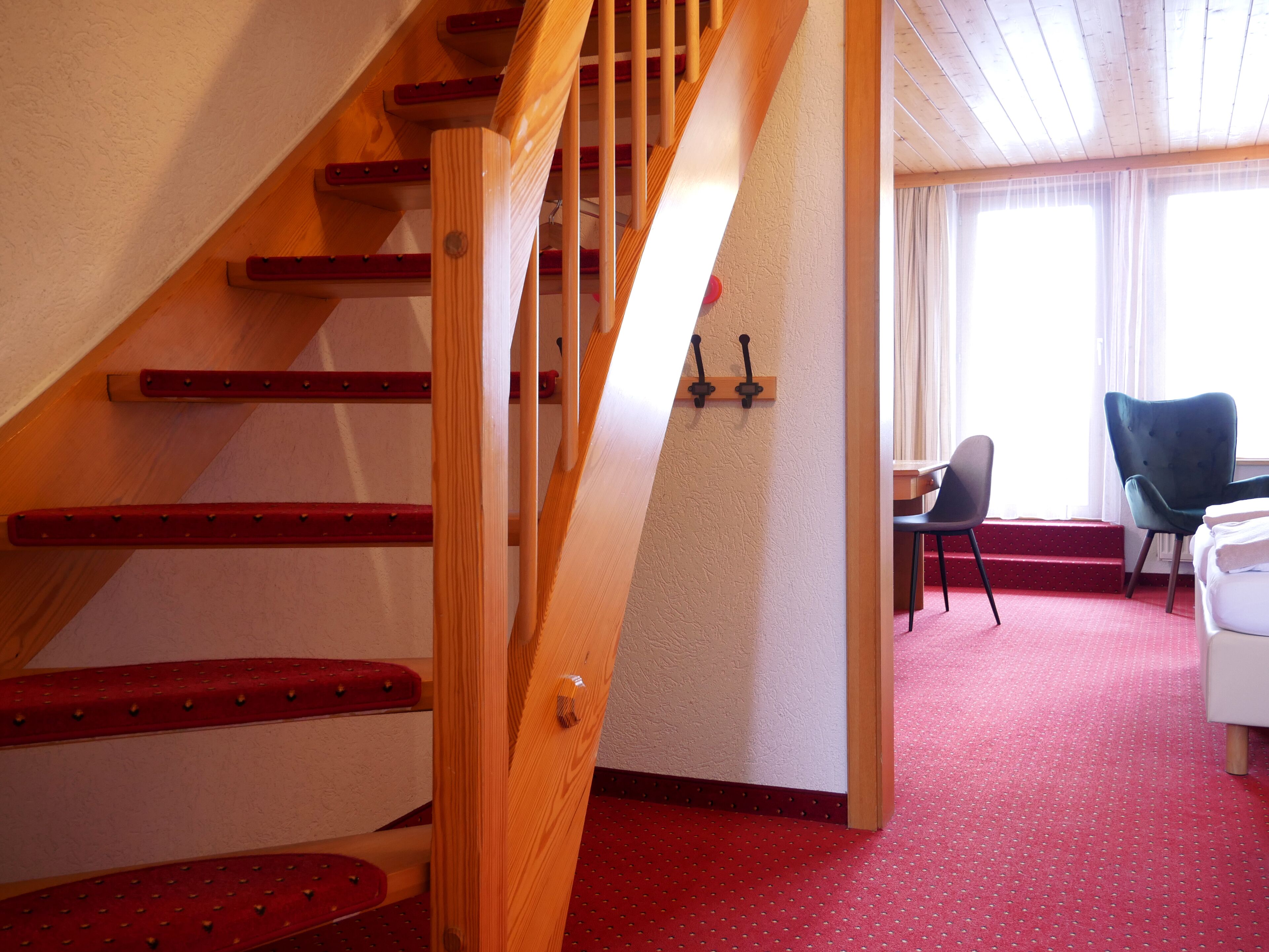 A staircase with wooden steps and red accents, leading to a room with a red carpet, a desk, and two chairs.