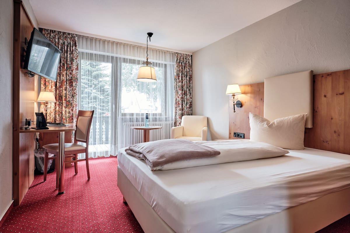 Habitación de hotel con cama, escritorio con silla, televisor, lámpara y ventana con cortinas.