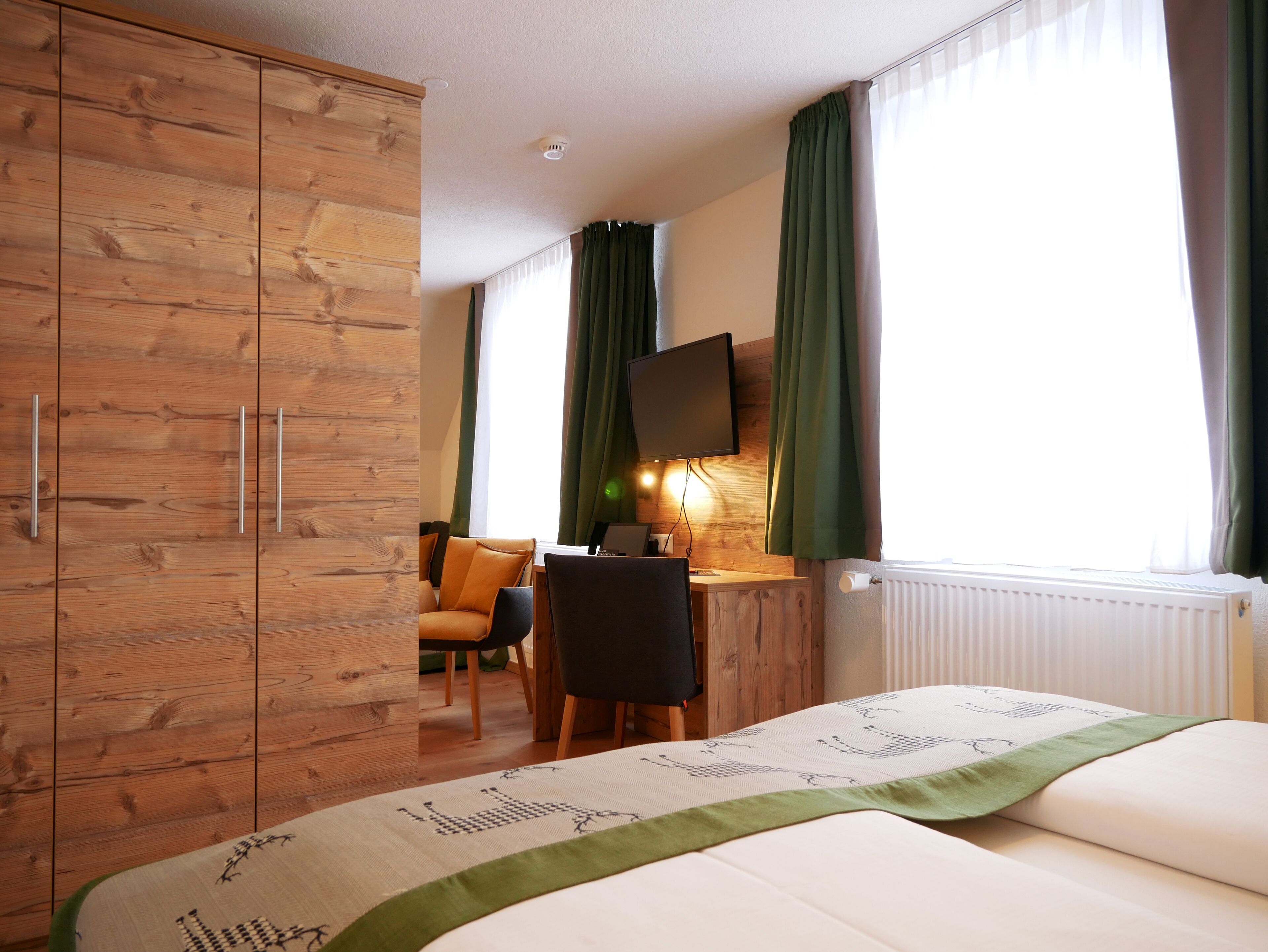 A hotel room with a wooden wardrobe, a bed with a green accent, a desk with a chair, a television mounted on the wall, and a window with curtains.