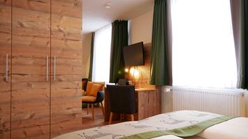 A hotel room with a wooden wardrobe, a bed with a green accent, a desk with a chair, a television mounted on the wall, and a window with curtains.