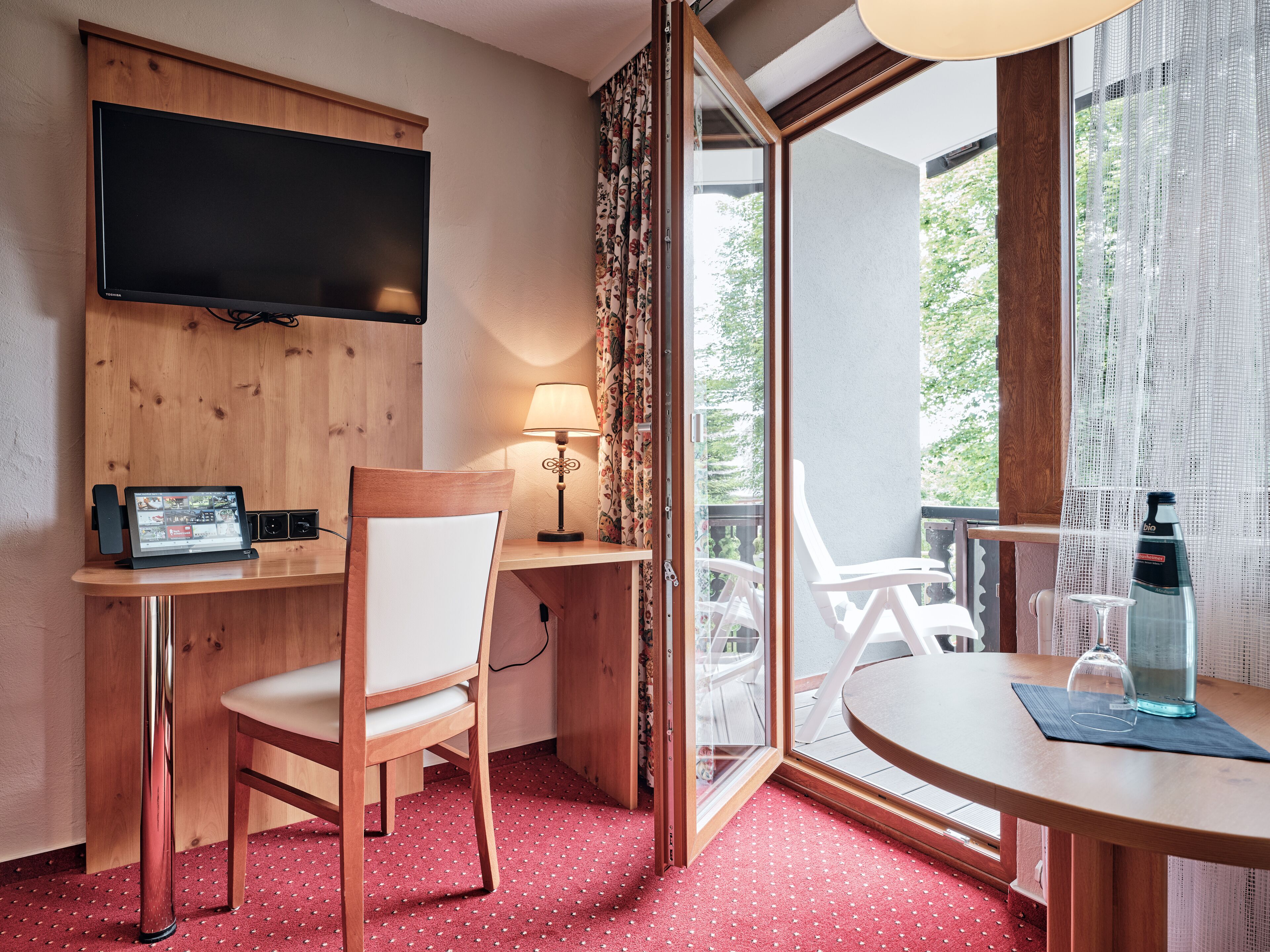 A hotel room with a wooden desk, a chair, a TV mounted on the wall, a table with a bottle and glass, and a view of greenery through the window.