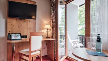 Quarto de hotel com escrivaninha de madeira, uma cadeira, TV fixada na parede, uma mesa com garrafa e copo, e vista para o verde pela janela.