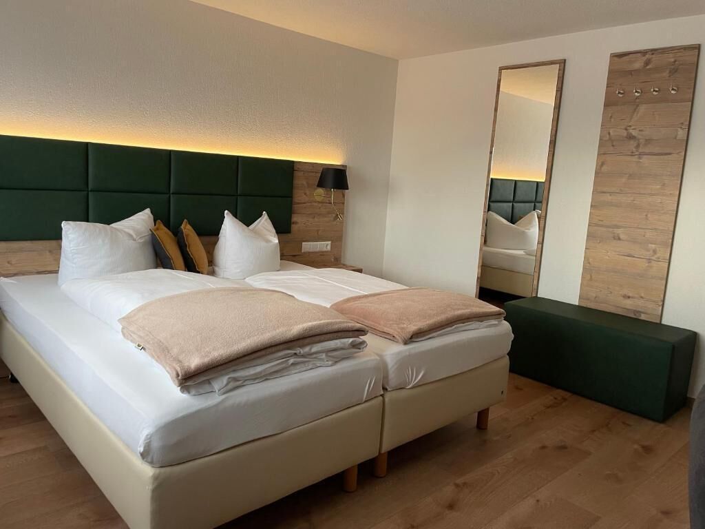A double bed with white and beige linens, a green upholstered headboard, a bedside table with a lamp, a large mirror, and a green bench.