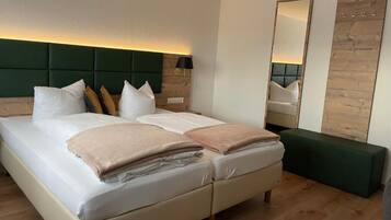 A double bed with white and beige linens, a green upholstered headboard, a bedside table with a lamp, a large mirror, and a green bench.