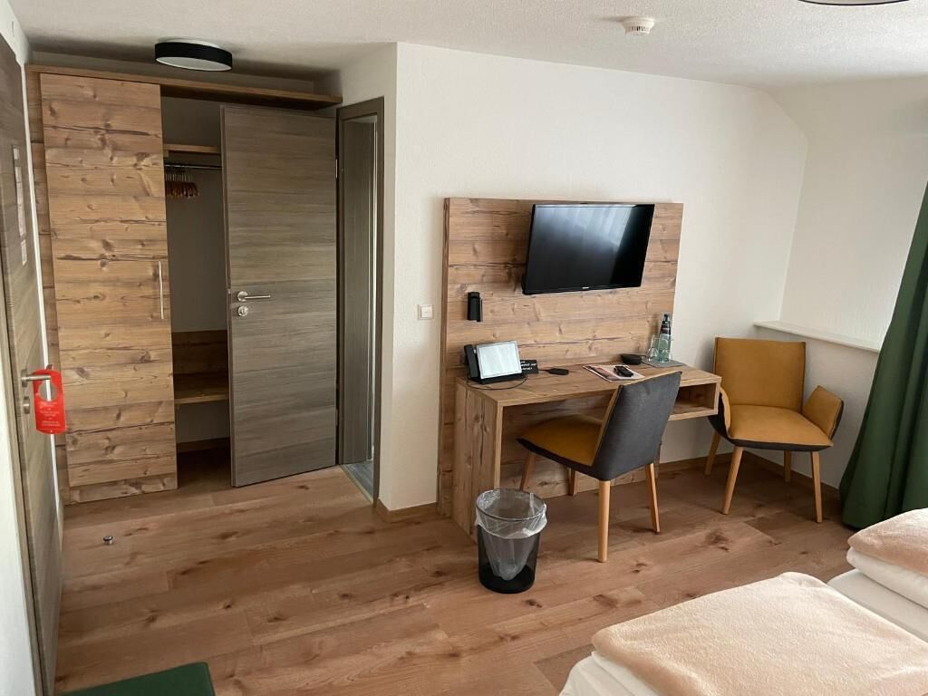 A hotel room with a wooden desk, a television mounted on the wall, a chair, a bed, and a wardrobe.