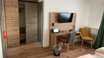 A hotel room with a wooden desk, a television mounted on the wall, a chair, a bed, and a wardrobe.