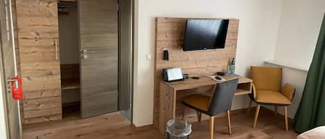 A hotel room with a wooden desk, a television mounted on the wall, a chair, a bed, and a wardrobe.