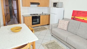 Un espacio habitable compacto con kitchenette, una mesa de comedor con sillas, un sofá con un cojín estampado y un cuadro en la pared.