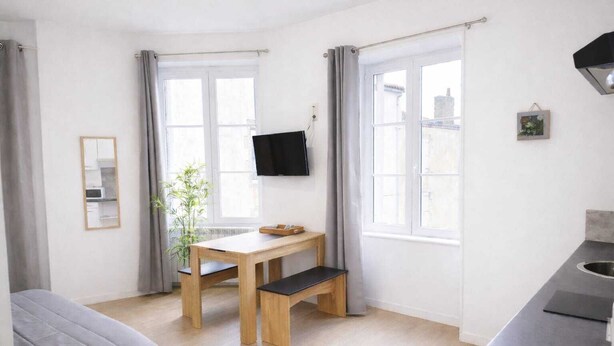 Una habitación pequeña y luminosa con una cama, una mesa con una silla, un televisor y una ventana.