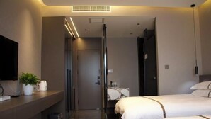 Ein modernes Hotelzimmer mit einem großen Bett, einem Nachttisch mit Pflanze, einem Flachbildfernseher und einem durch eine Glastür einsehbaren Badezimmer.