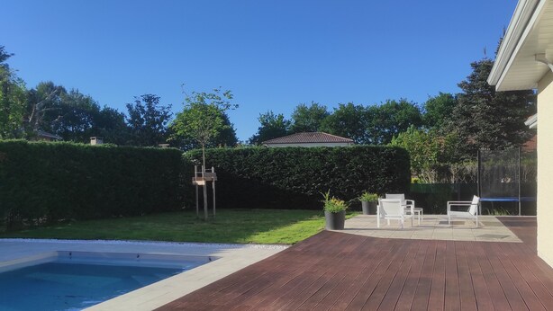 Een houten terras met een zwembad, tuinmeubilair en een verzorgde tuin.