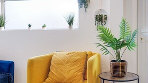 A yellow armchair with a herringbone pillow, a round wooden side table with a potted plant, and a blue chair.