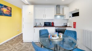 A modern kitchen with white cabinets, a microwave, and a kettle. There is a glass table with blue chairs and a bowl of cereal.
