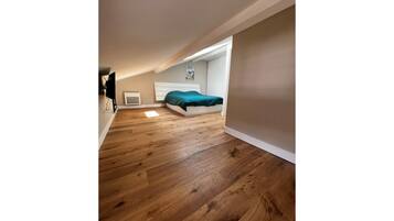 Une chambre avec un plancher en bois, un lit recouvert d’une couverture verte, une tête de lit blanche et une petite fenêtre.