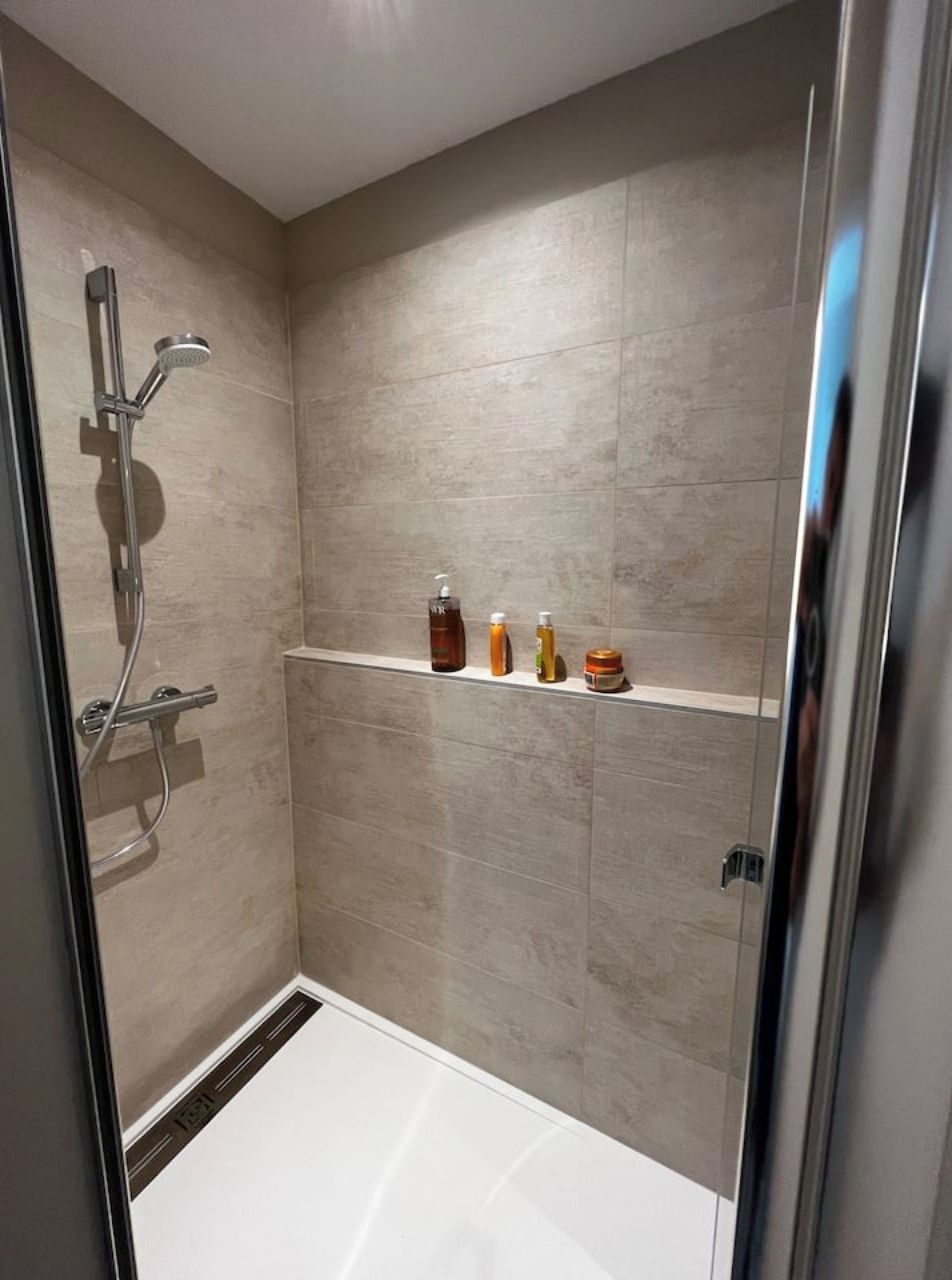 A modern shower with a glass door, a handheld showerhead, and a shelf with toiletries.