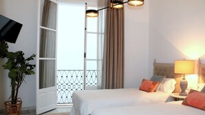 A hotel room with two beds, a TV mounted on the wall, a balcony with a metal railing, and a lamp on a bedside table. - Hostal Roomsole (Seville)