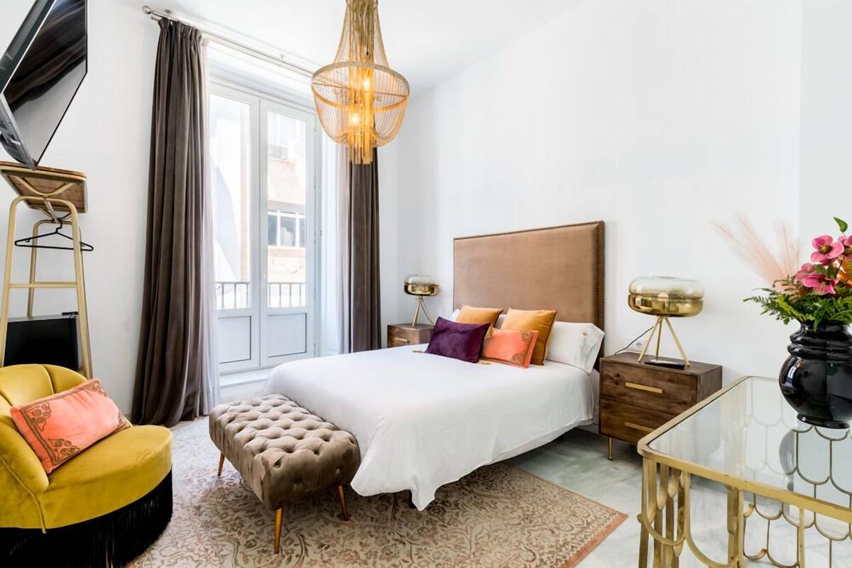 A bedroom with a bed, bedside tables, a chair, a bench, a hanging light fixture, and a window.