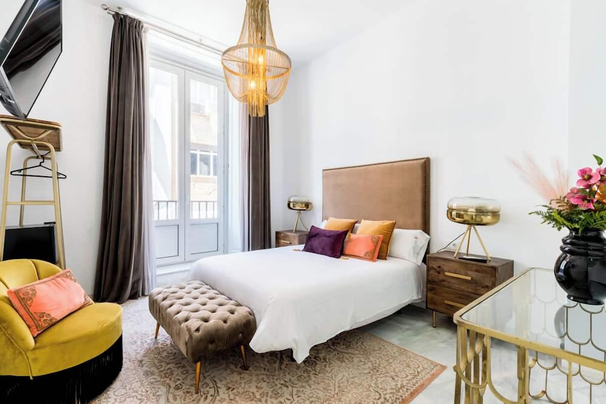 A modern hotel room with a large bed, a sitting area with a yellow chair, a glass coffee table, and a decorative chandelier.