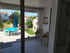 Quarto com porta de correr de vidro que dá para um pátio. O pátio possui uma mesa com cadeiras e toalha de mesa azul. Há duas cadeiras dentro do quarto e um adorno pendurado na parede.