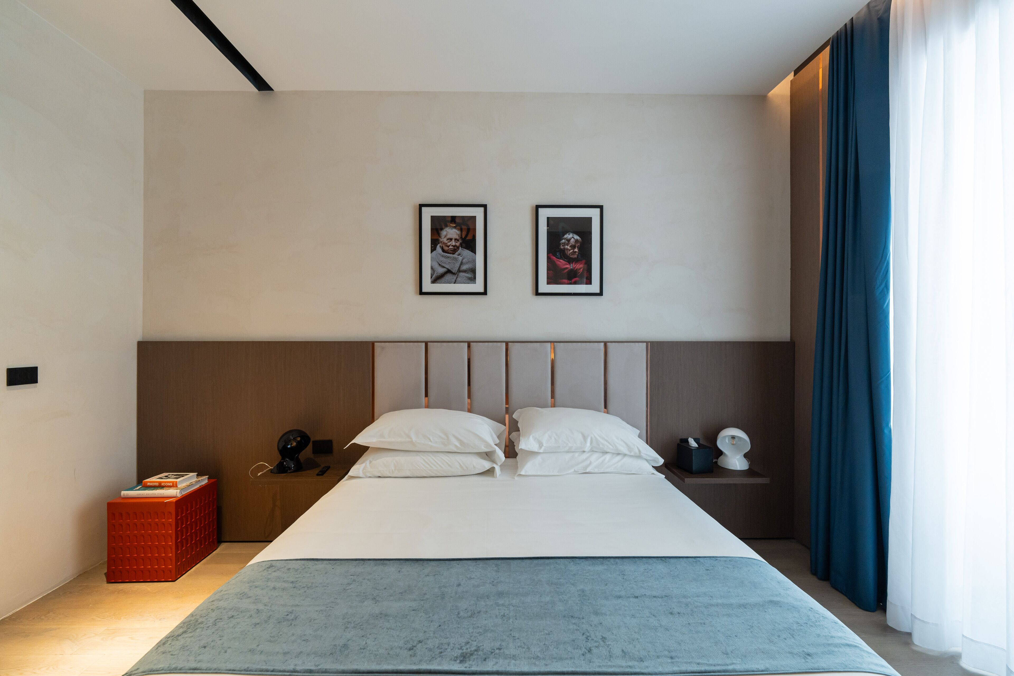 Un dormitorio moderno con una cama grande, dos cuadros enmarcados en la pared, una mesita de noche con una lámpara y un caja roja de almacenamiento.