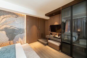 A modern bedroom with a large bed, a TV, a wardrobe, and a bathroom visible through a glass door. - Agra Suites (Naples)
