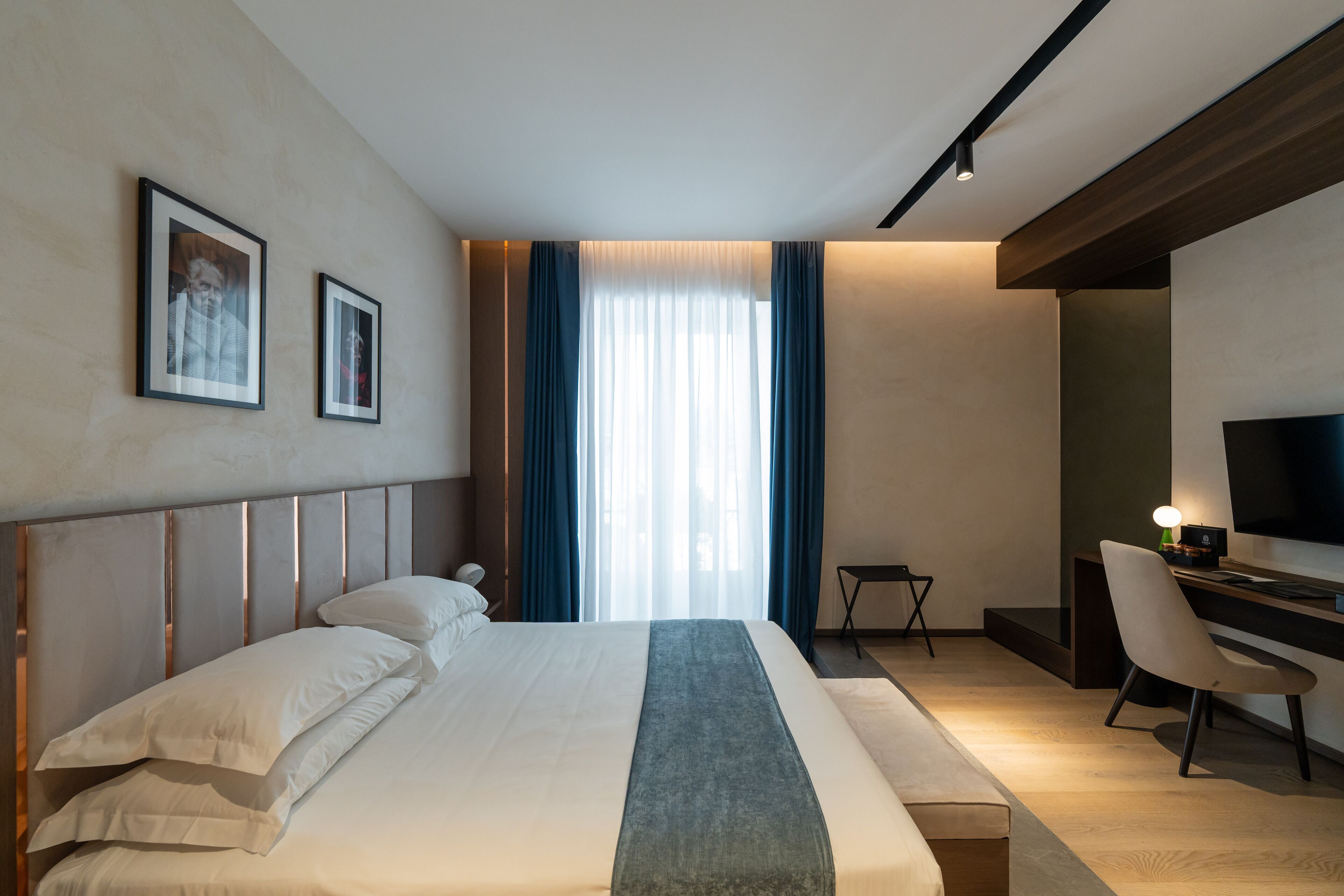 A modern hotel room with a large bed, a desk with a chair, a television, and two framed pictures on the wall.