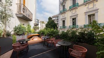 A rooftop patio with wicker furniture, a small tree, and a view of adjacent buildings.