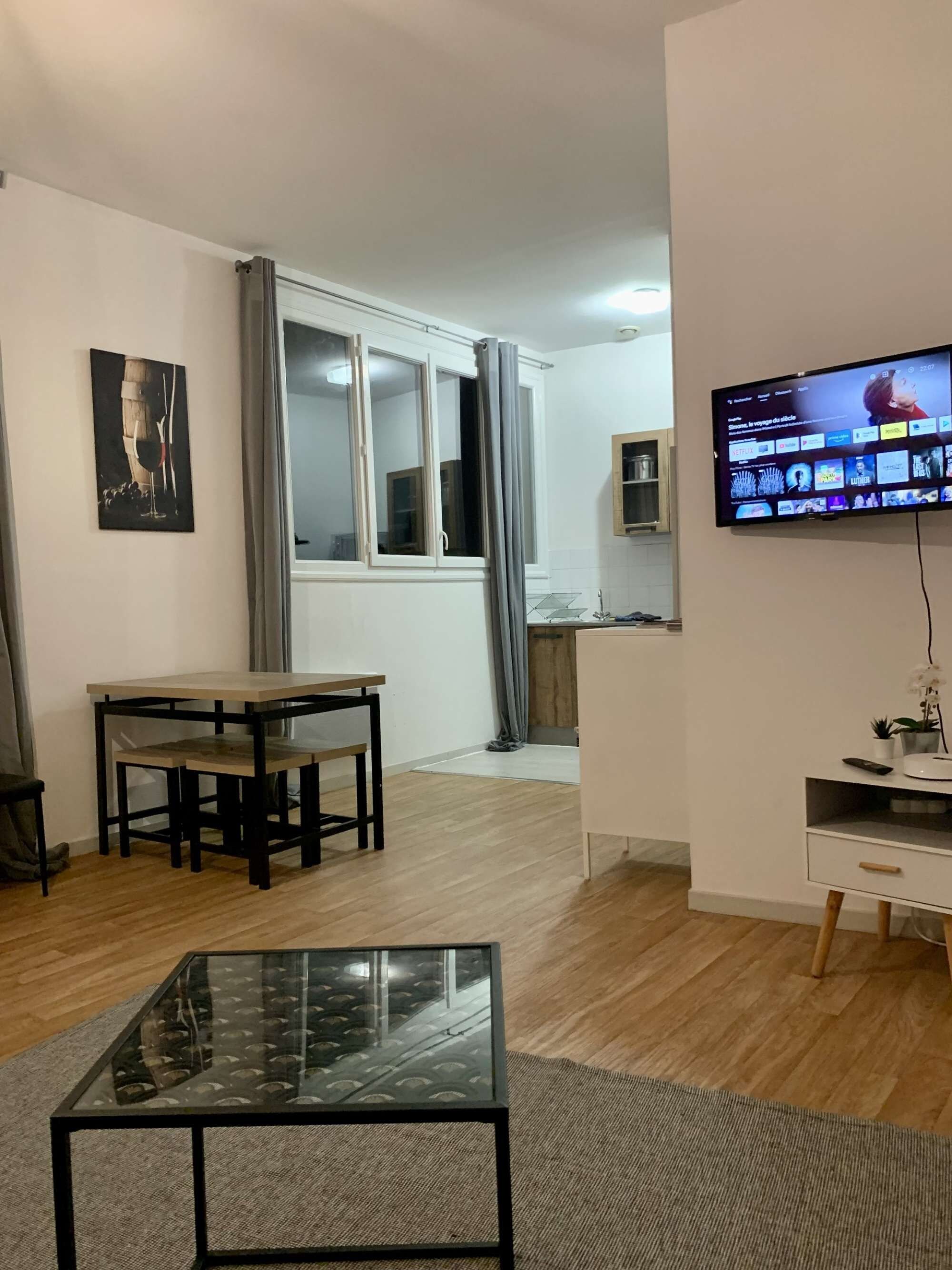 Un salon avec une table basse en verre, une table à manger avec des chaises, un téléviseur fixé au mur et, au fond, un coin cuisine.