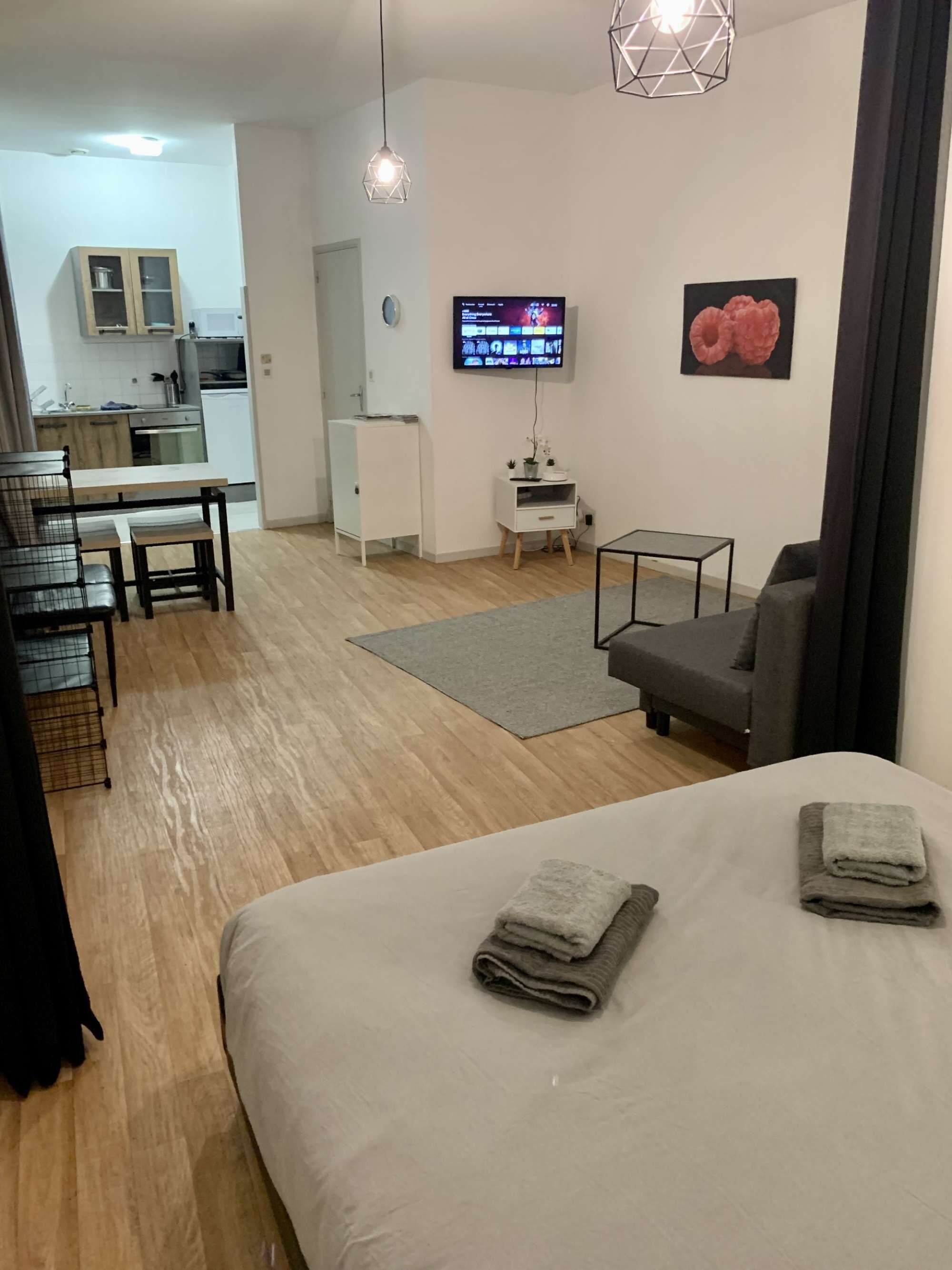Una habitación moderna con cama, una pequeña mesa, un sofá gris, televisión y una cocina al fondo.