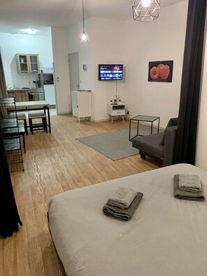 A modern room with a bed, a small table, a grey sofa, a TV, and a kitchen in the background.