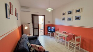 Een woonkamer met een bank, een televisie, een tafel met stoelen en een deur naar een buitenruimte.